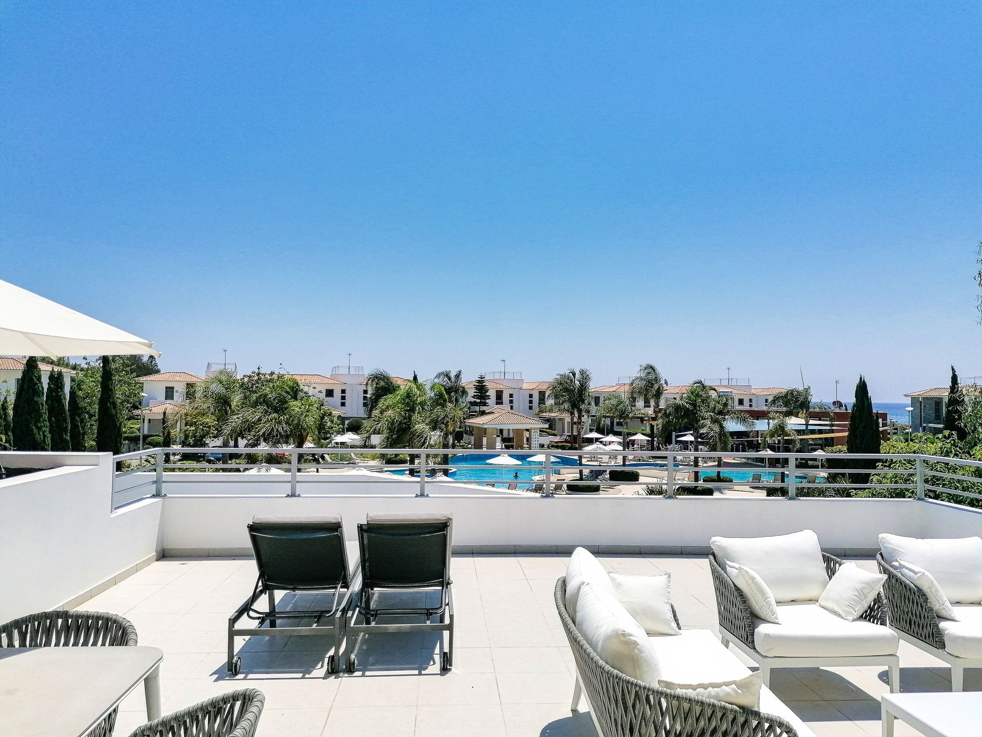 Rooftop terrace with lounge chairs, view of a pool, buildings, and blue sky.