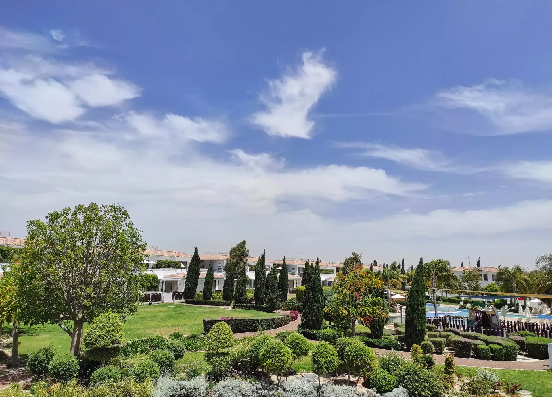 Lush green landscape with trees, shrubs, and houses under a partly cloudy, bright blue sky.