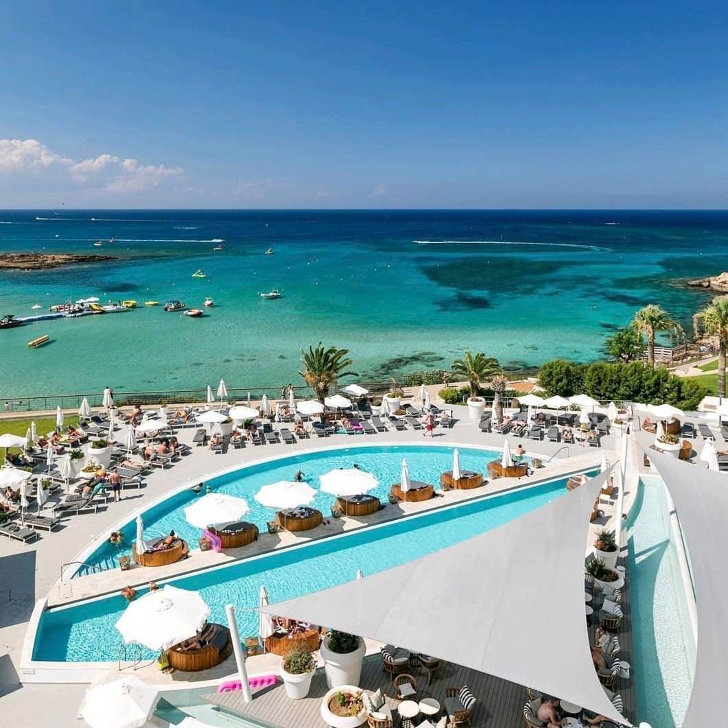 King Jason Protaras hotel overlooking turquoise water; umbrellas, loungers, and boats visible.