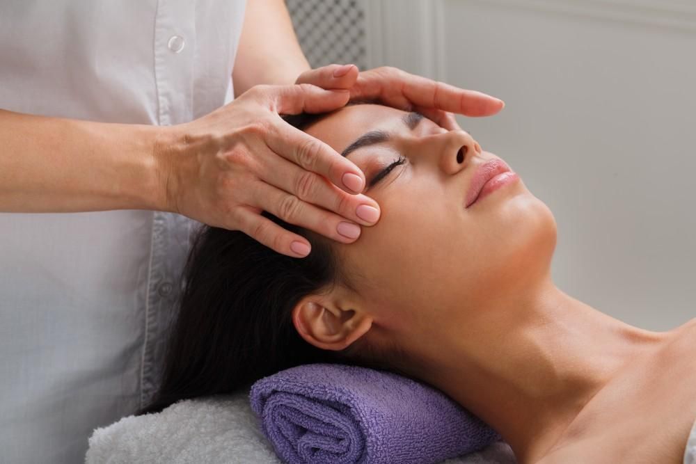 Hands gently massage a woman's forehead and temples as she reclines on a purple towel.
