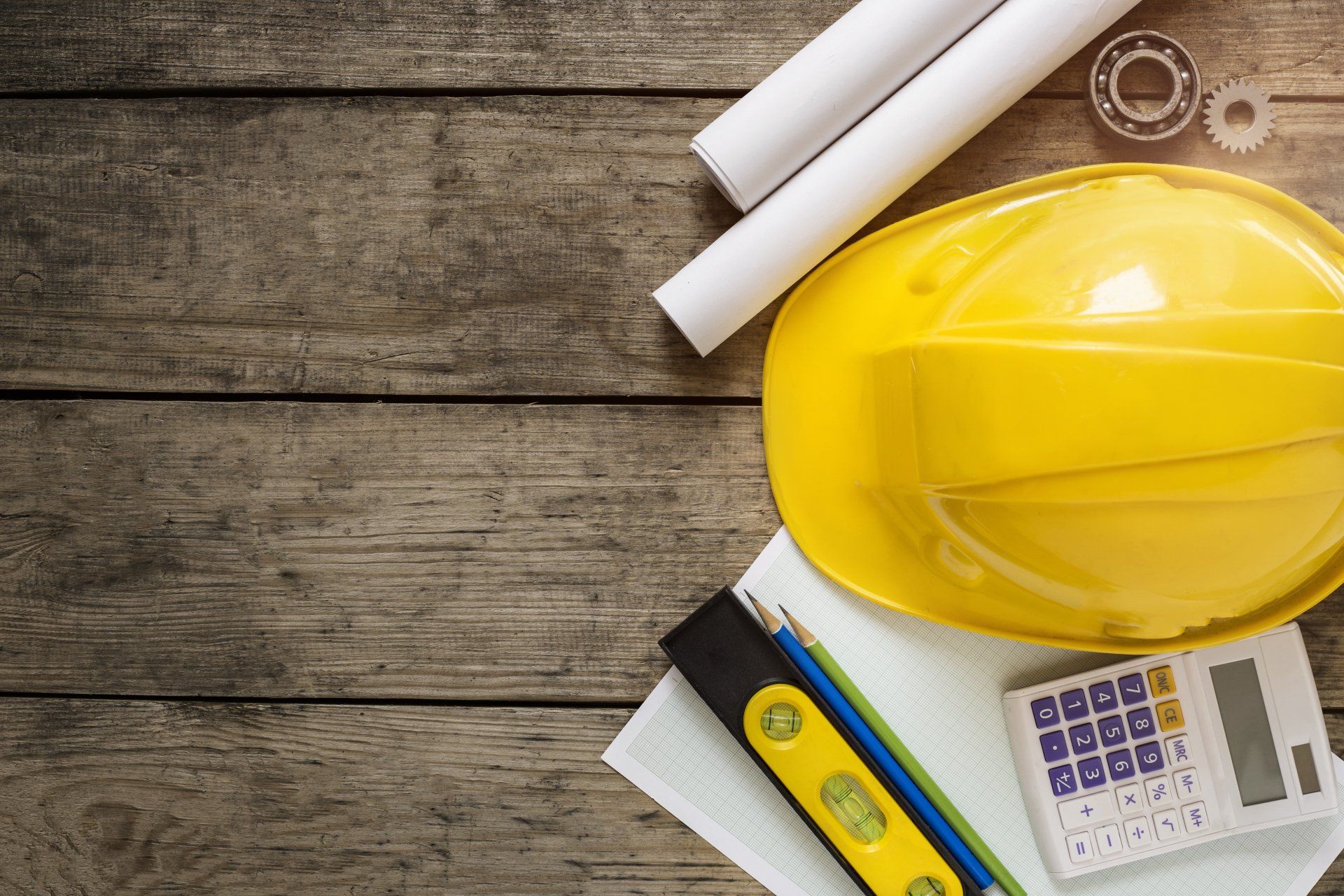 Auf einem Holztisch liegen ein gelber Schutzhelm, ein Taschenrechner, eine Wasserwaage und ein Bleistift.
