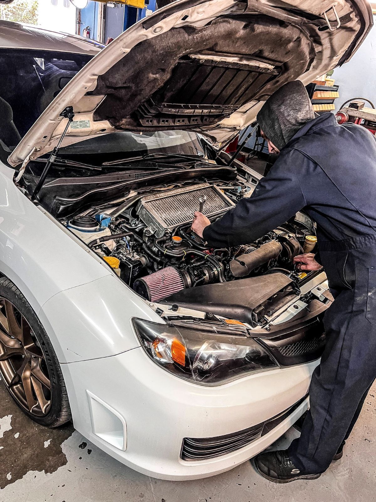 A mechanic in dark coveralls works on the engine of a white car with its hood open in a garage.