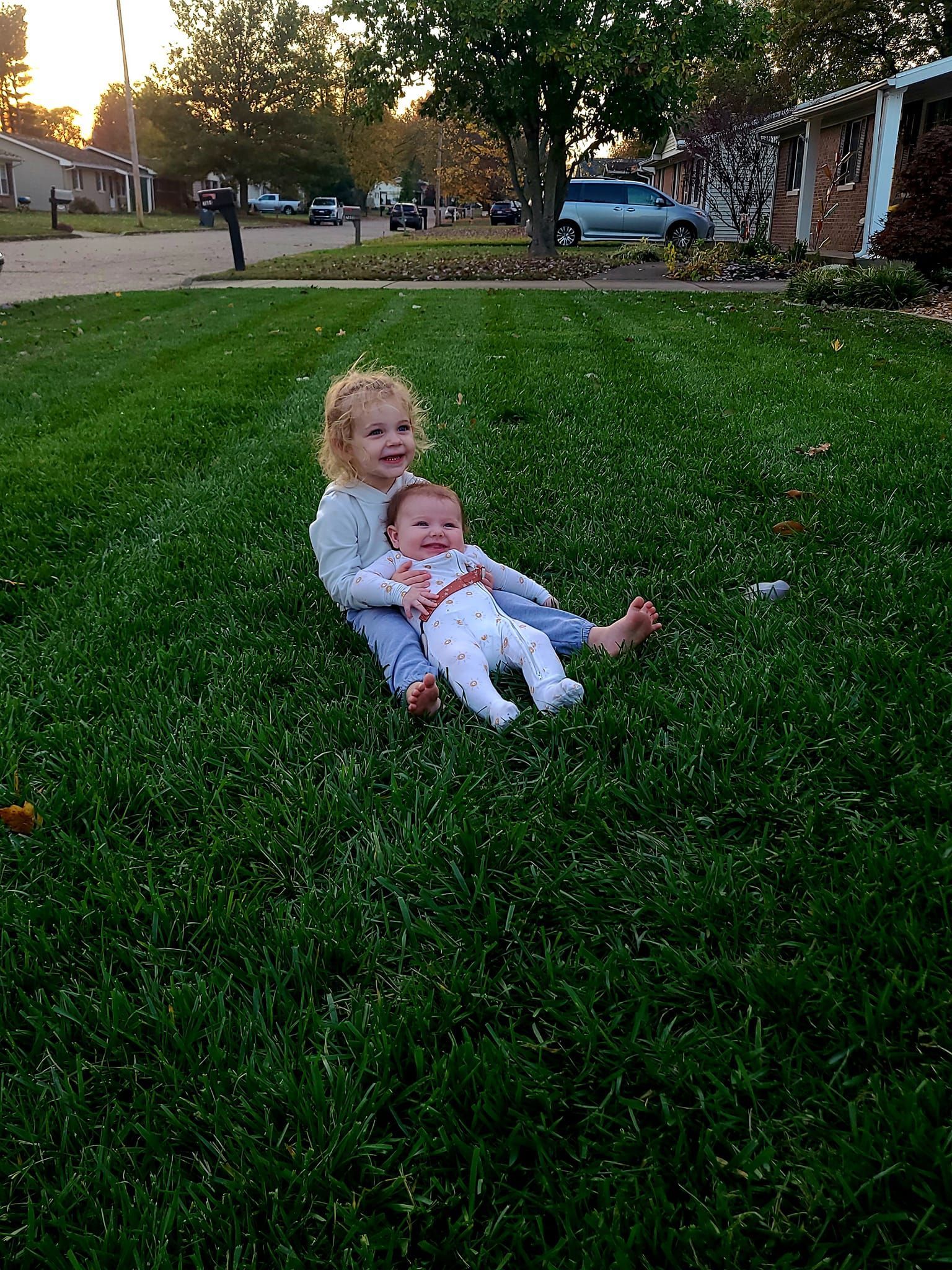 Two children sitting on green grass. One smiles, the other looks up. Houses and vehicles in the background.