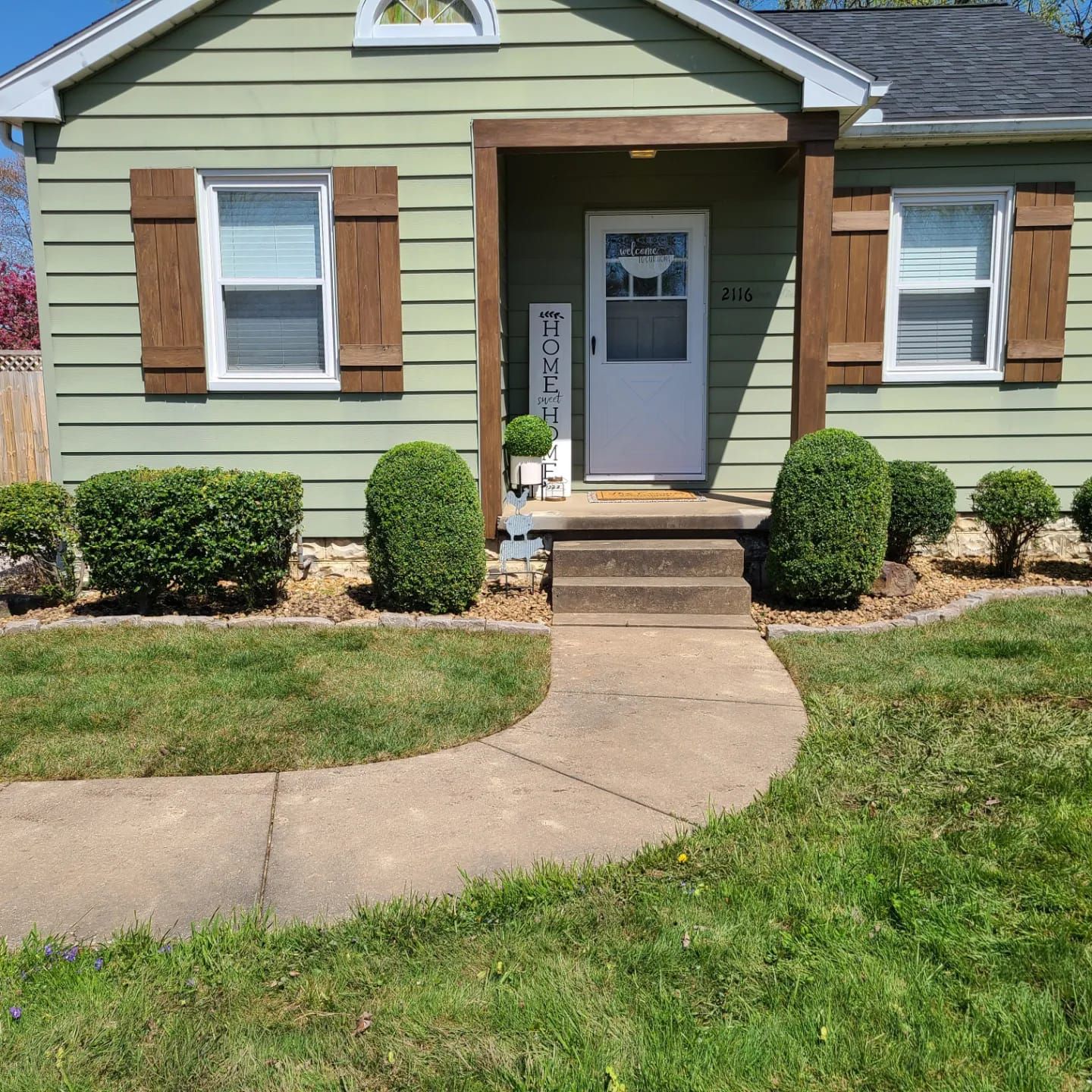 A light green house with brown shutters and a welcoming front porch, surrounded by green shrubs and a curved walkway.