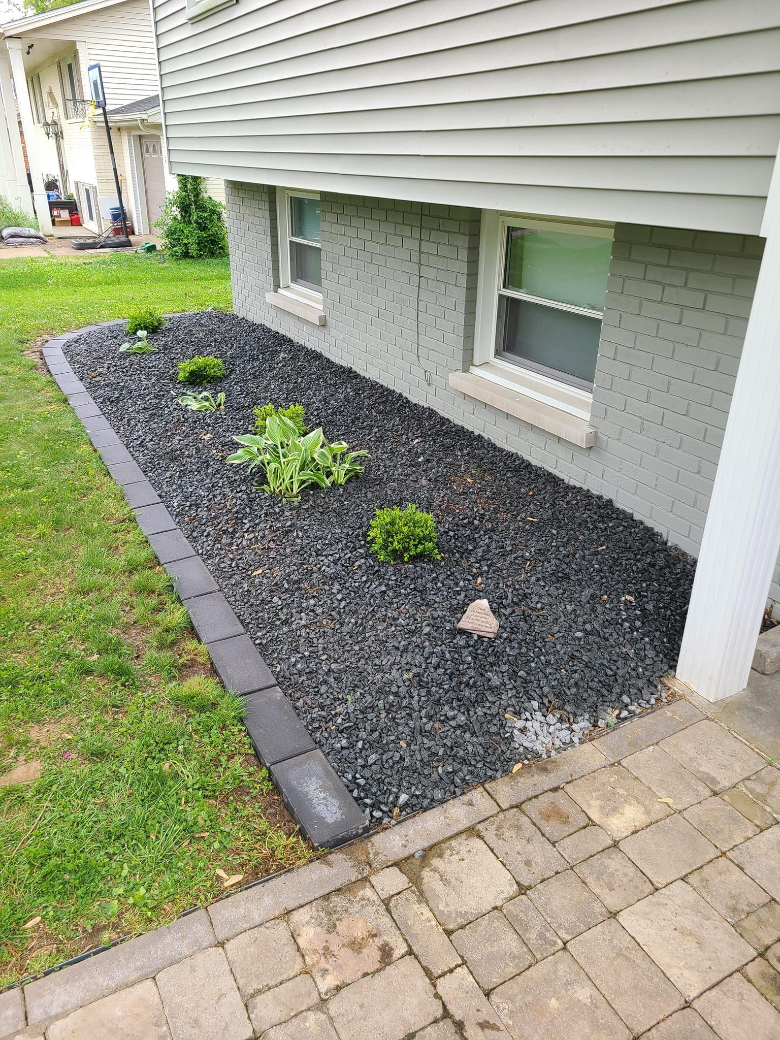 Black rock-filled landscaping bed with small plants and a dark border next to a light gray house.