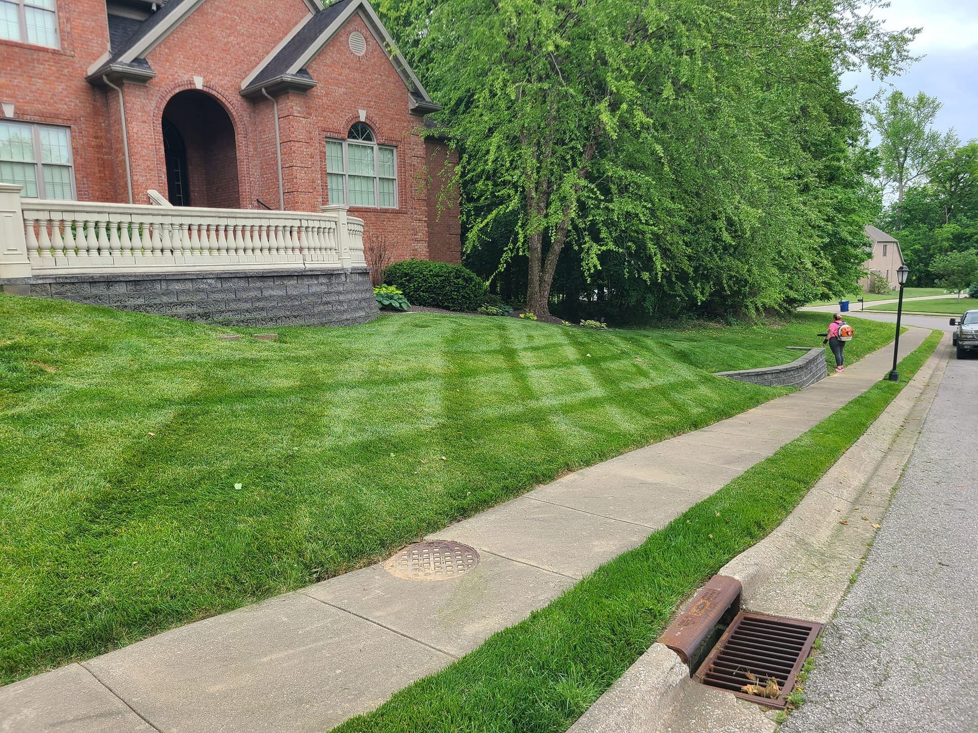Lawn with striped pattern next to a sidewalk and a brick house. Person walks on sidewalk.