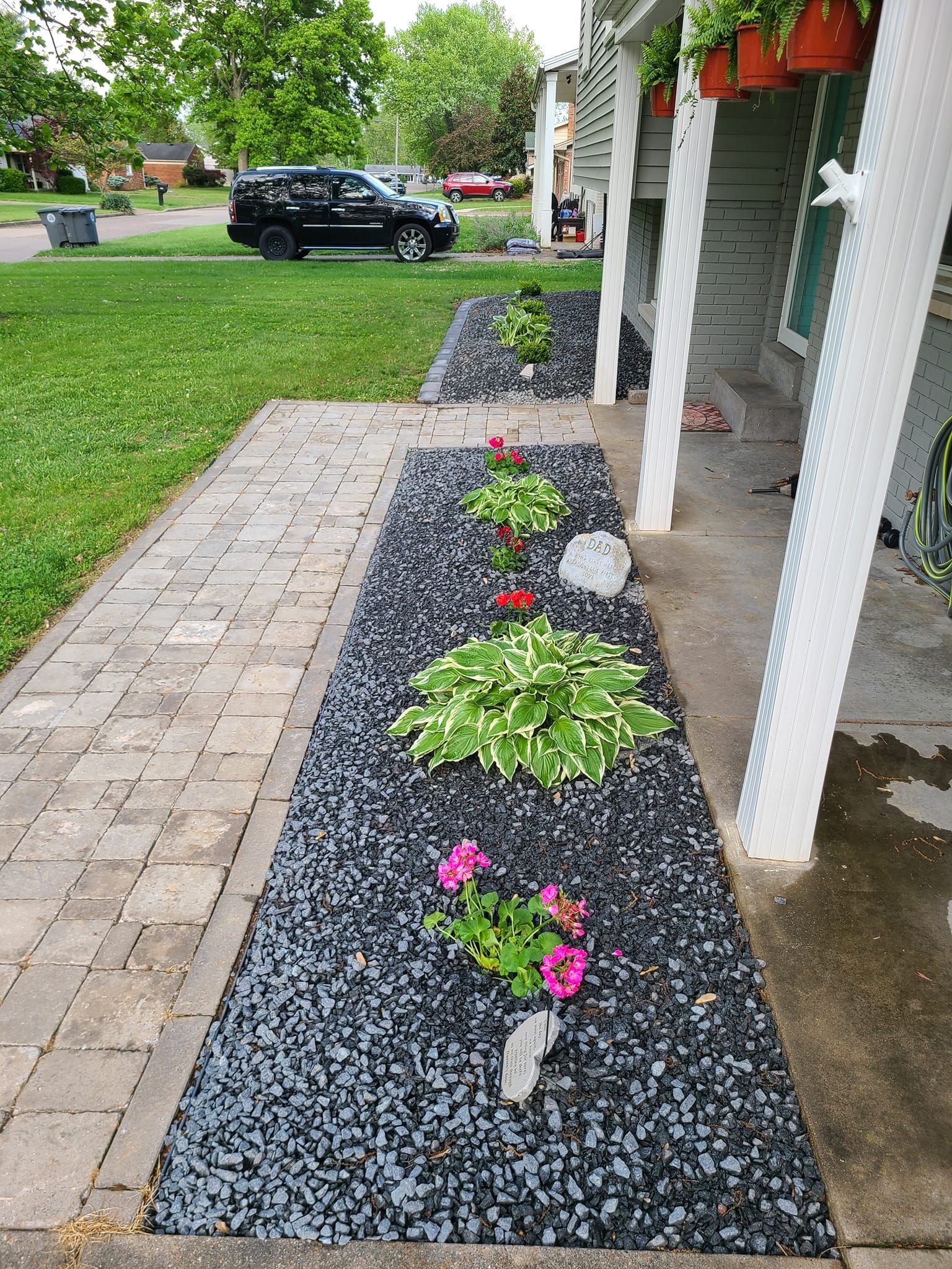 Black stone flowerbed with various plants along a paved walkway next to a house with a porch.