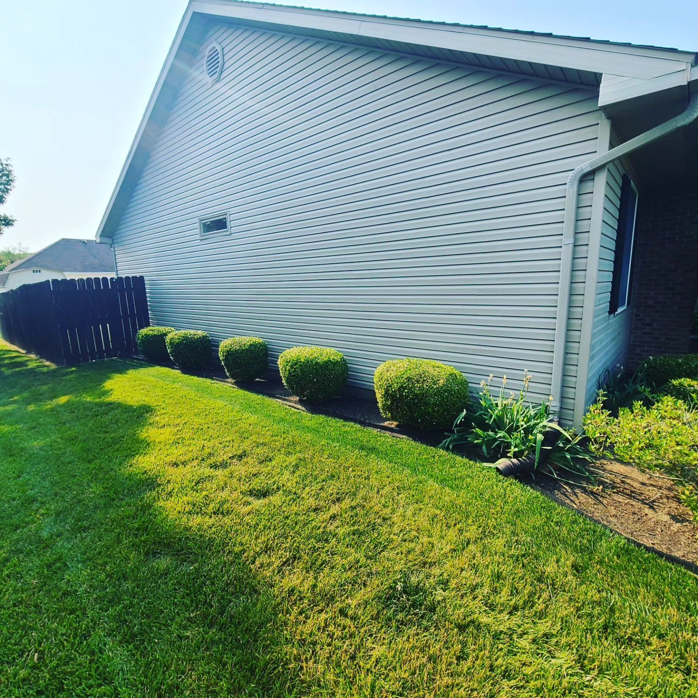 Side view of a house with light grey siding, green lawn, and neatly trimmed green bushes.