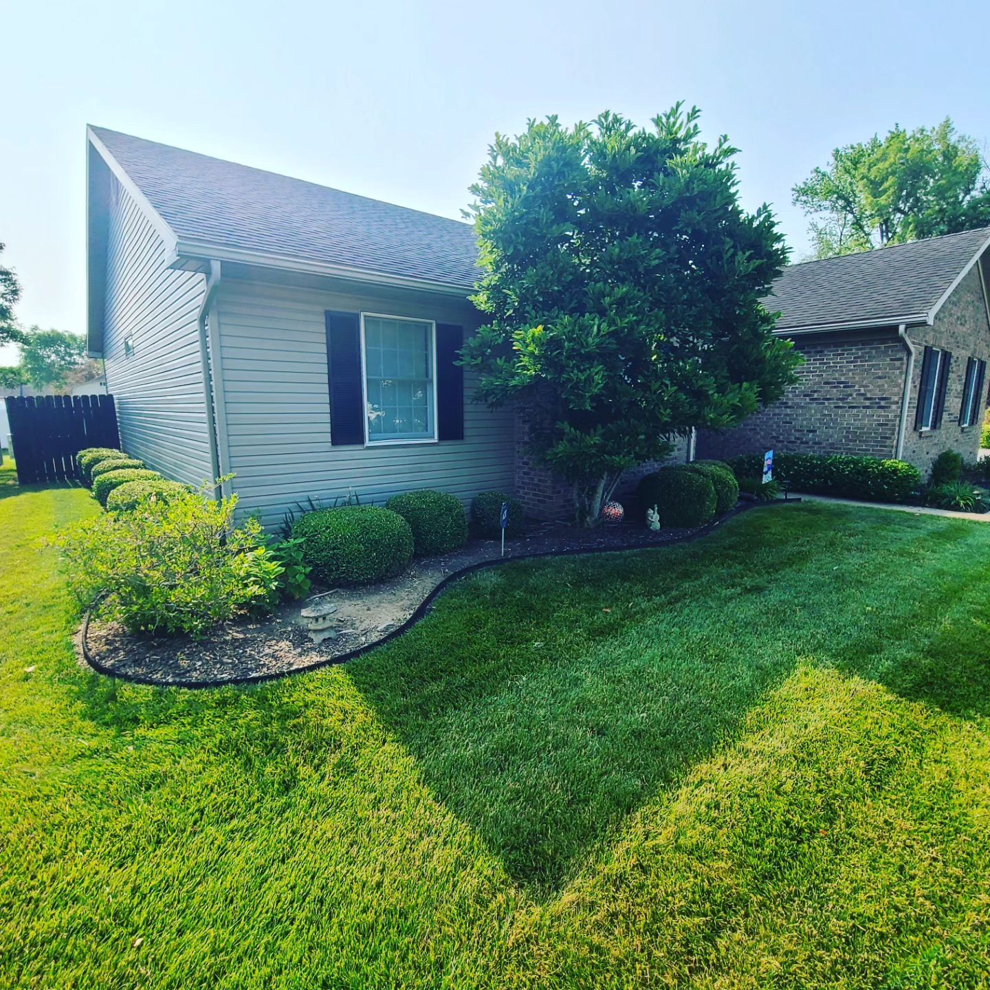 Well-manicured lawn and home with tan siding and dark shutters, blue sky.