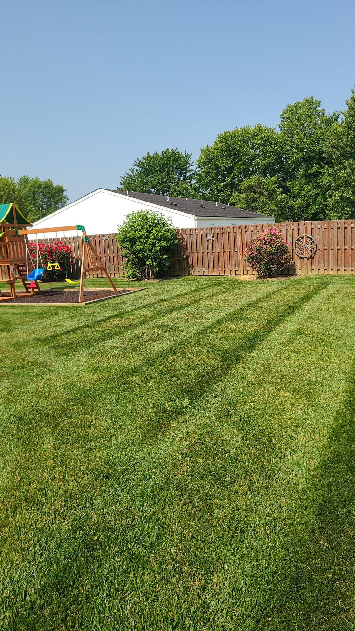 Lawn with stripes, swingset, fence, and a white-roofed building under a clear blue sky.