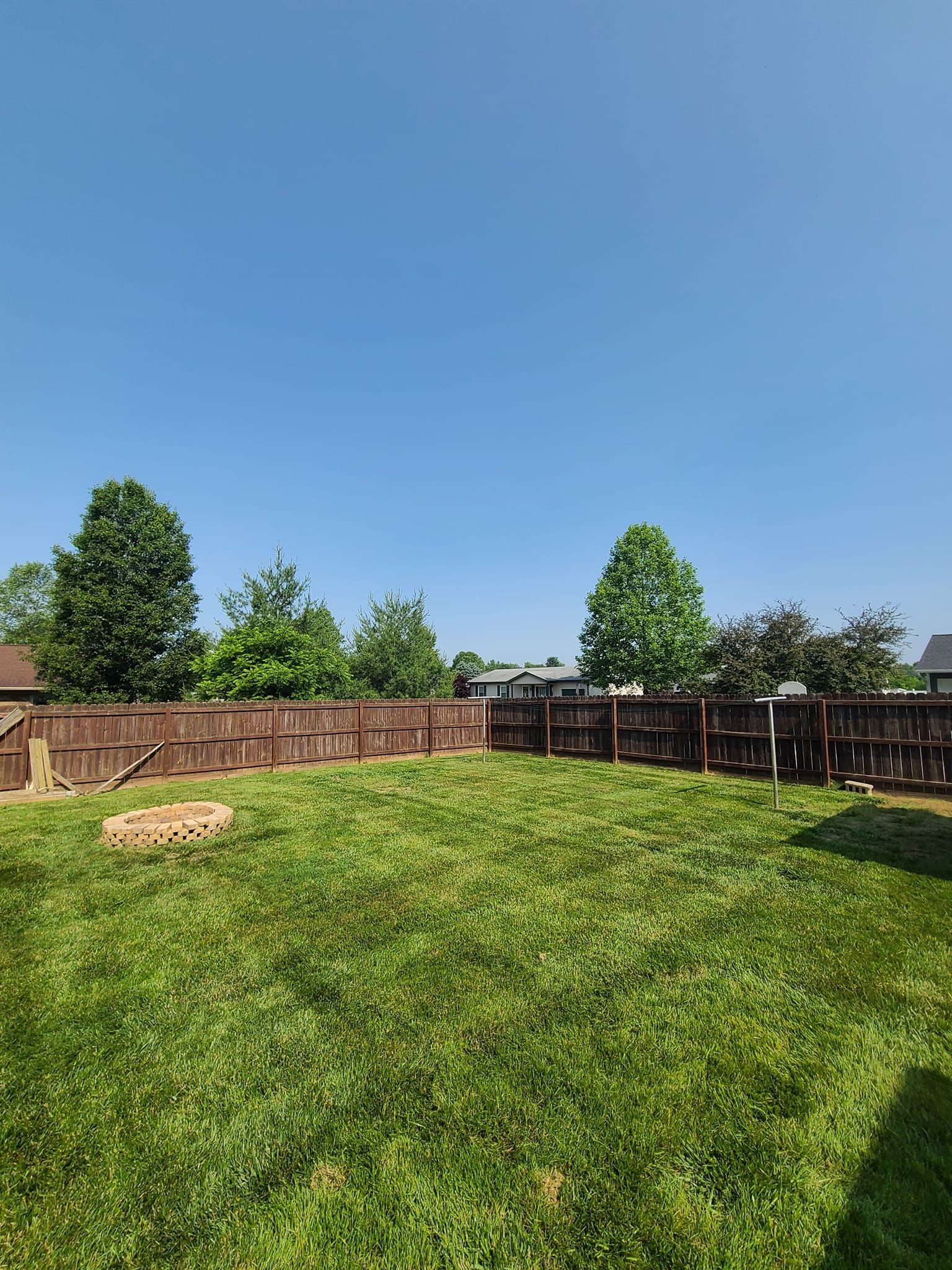 Green backyard with wooden fence, fire pit, trees, and blue sky.