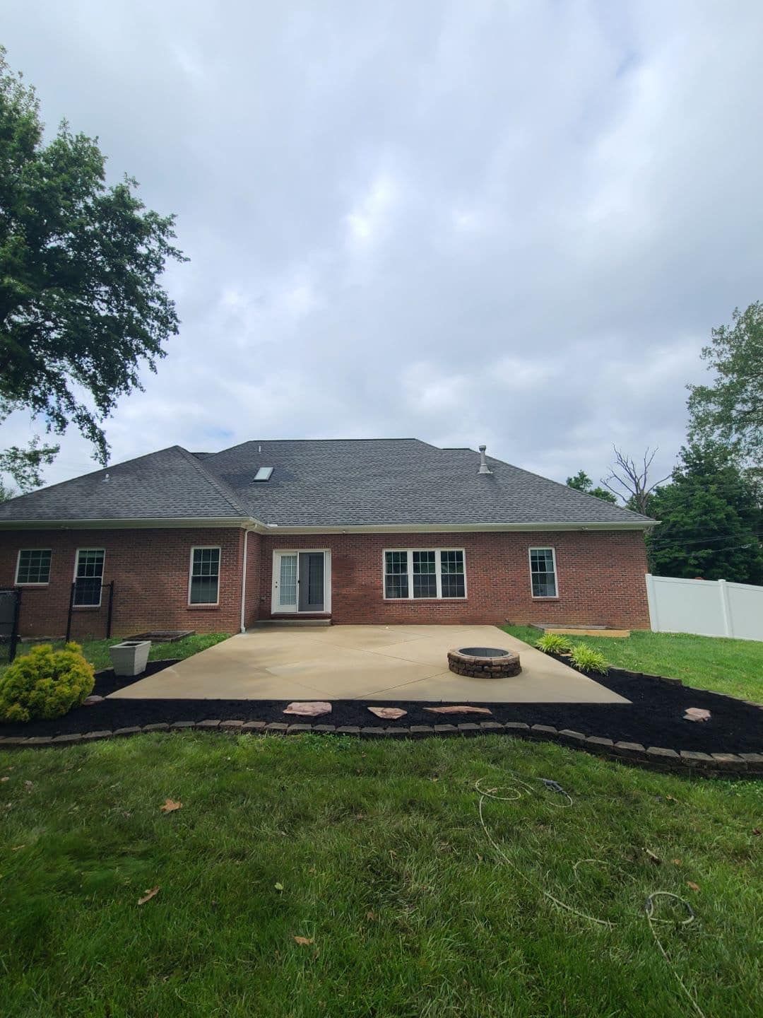 Back of a red brick house with a concrete patio and a fire pit, surrounded by green grass and mulch. Cloudy sky.
