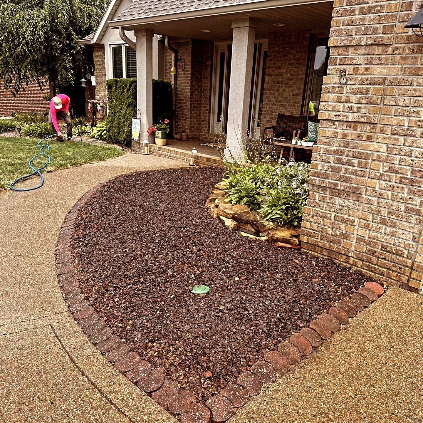 Curved walkway with flower bed bordered by brick, next to a brick house. Person gardening in the background.