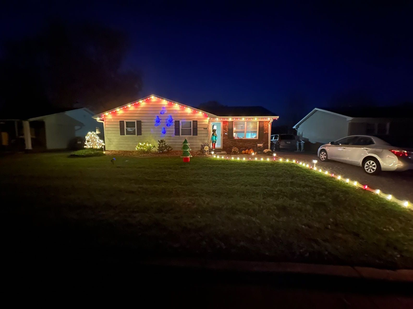 A house lit with Christmas lights at night, with a string of lights leading to a car in the driveway.