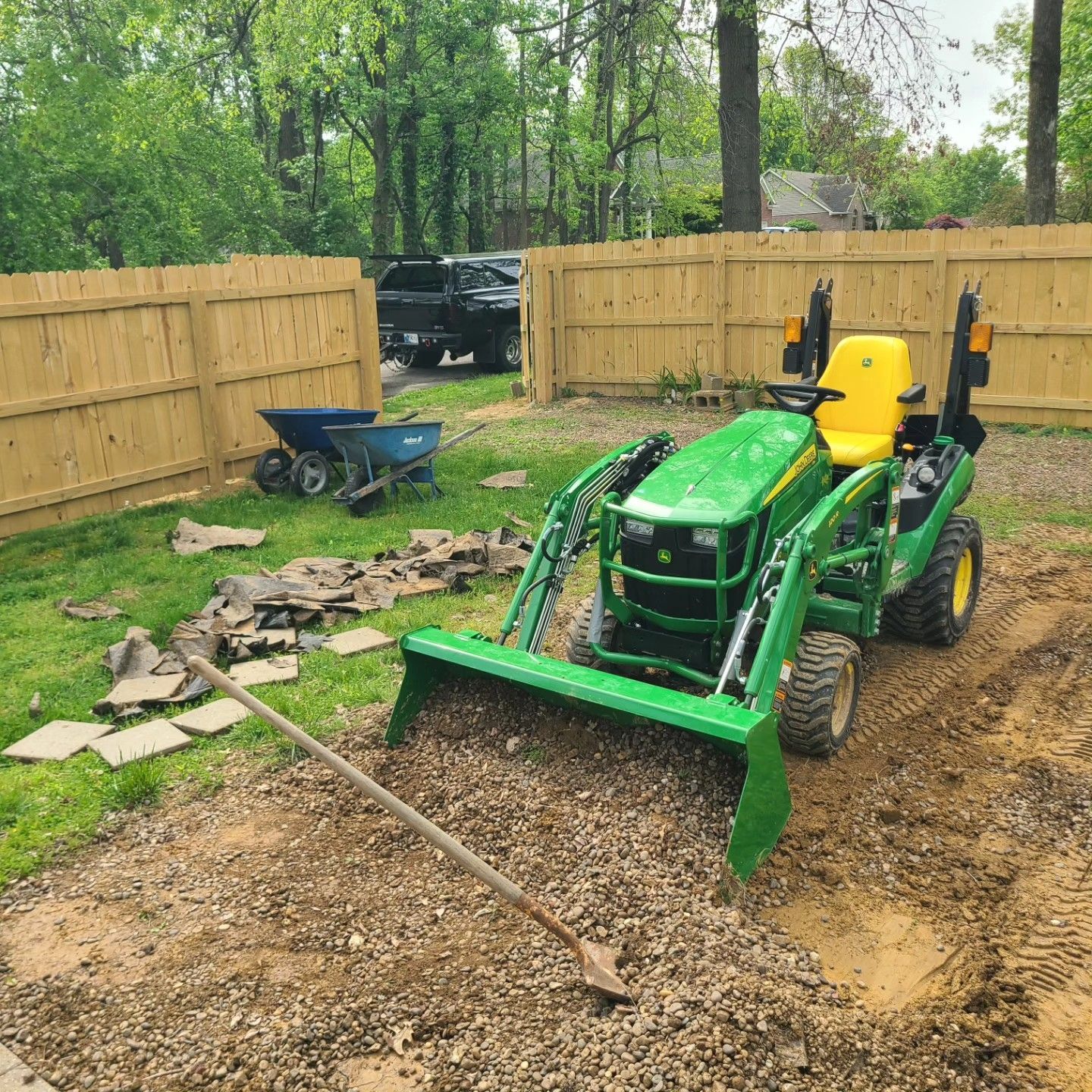 Green John Deere tractor moving gravel in a yard next to a wooden fence. A blue wheelbarrow and shovel are nearby.