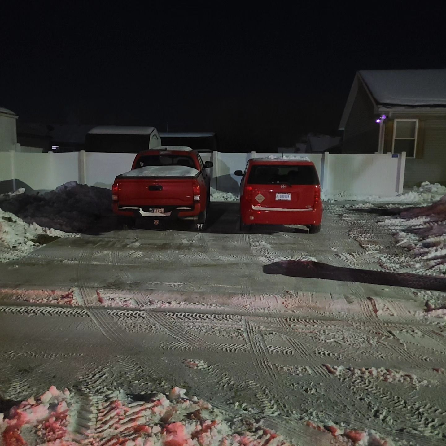 Two red vehicles parked in a snow-covered driveway at night. A white fence and house are in the background.