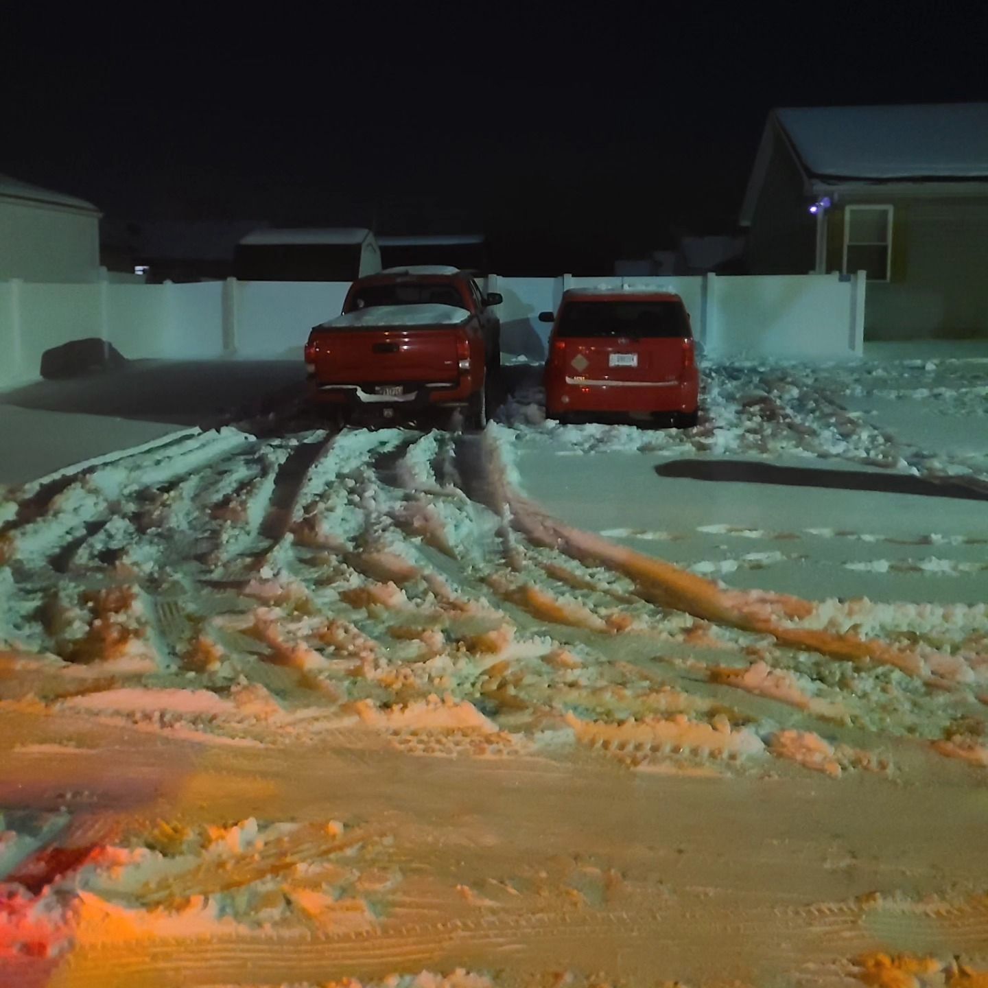 Red pickup truck and minivan parked in snow-covered driveway at night; tire tracks visible.