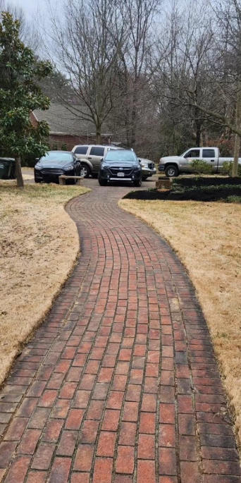 Brick pathway leading to cars parked near a house. Brown grass surrounds the path. Overcast day.
