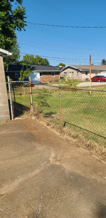 A weathered chain-link fence separates a cracked concrete area from a grassy yard with houses in the background.