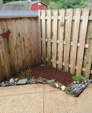 Corner garden bed with brown mulch, rocks, and green plants near a wooden fence.