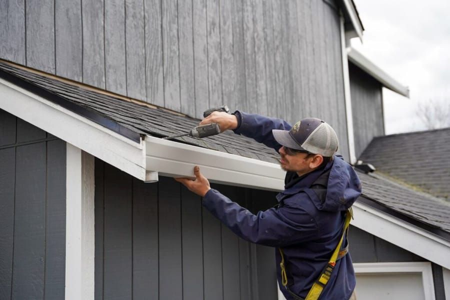 Person in blue jacket installs gutter on a gray house exterior, using a drill.