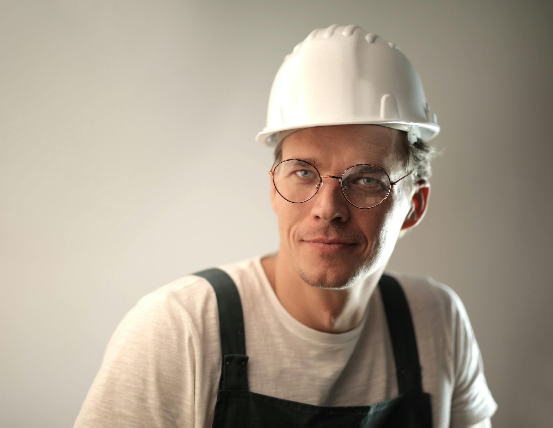 Man wearing a white hard hat and glasses, in work overalls, smiling.
