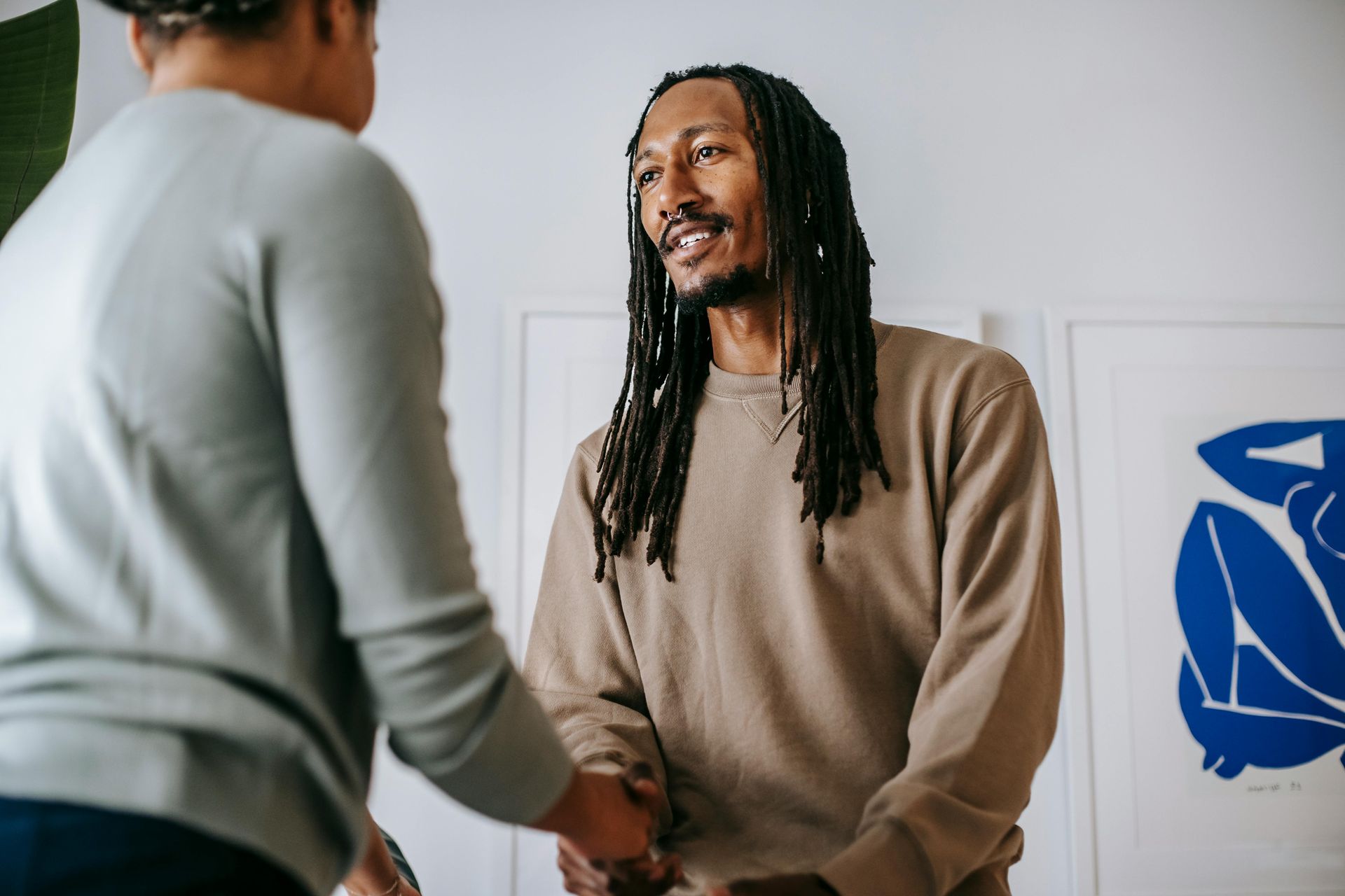 Man with dreadlocks smiling, shaking hands with another person indoors, art on the wall.