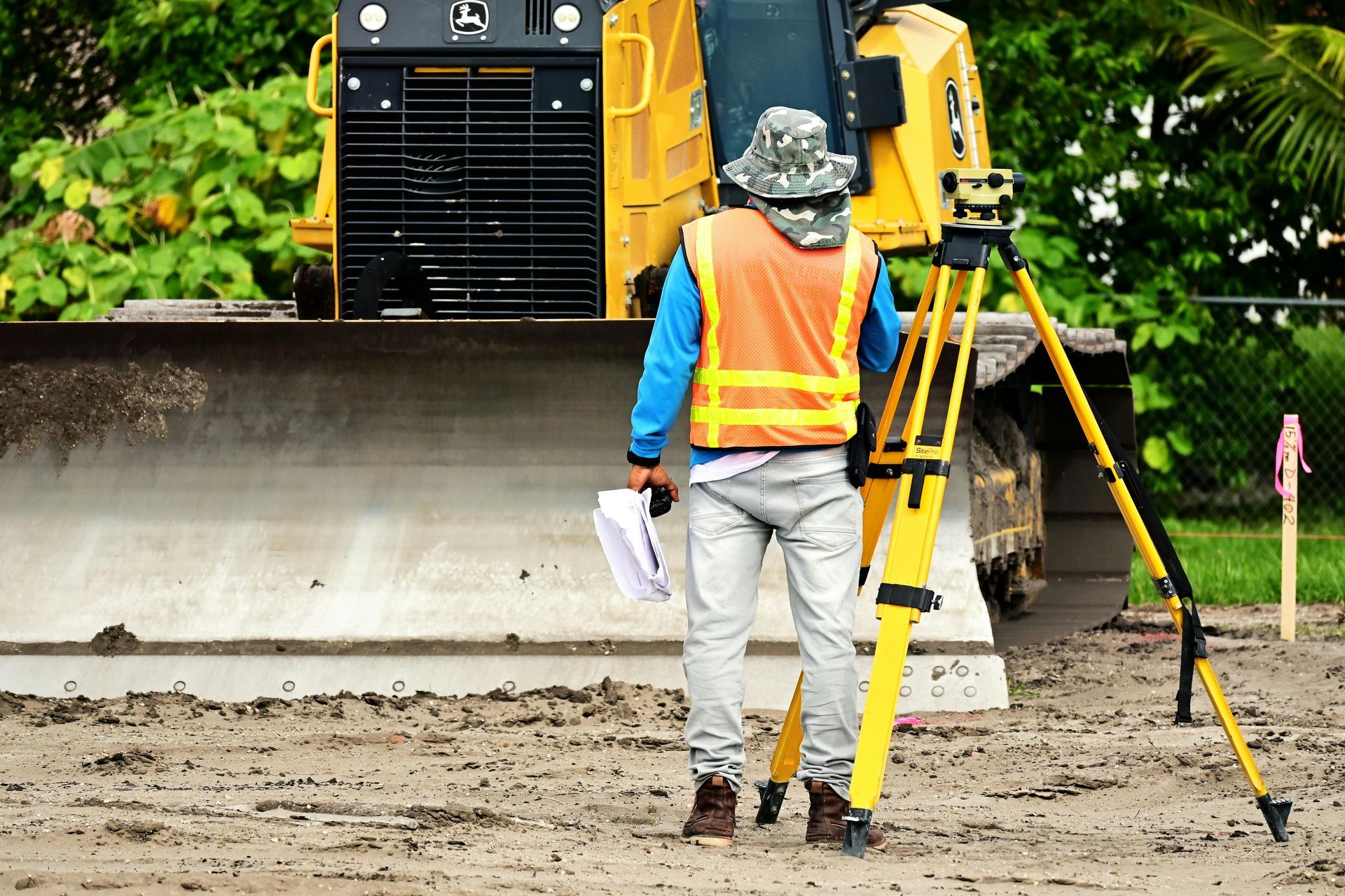 Surveyor with equipment at construction site, wearing safety vest, bulldozer in background.