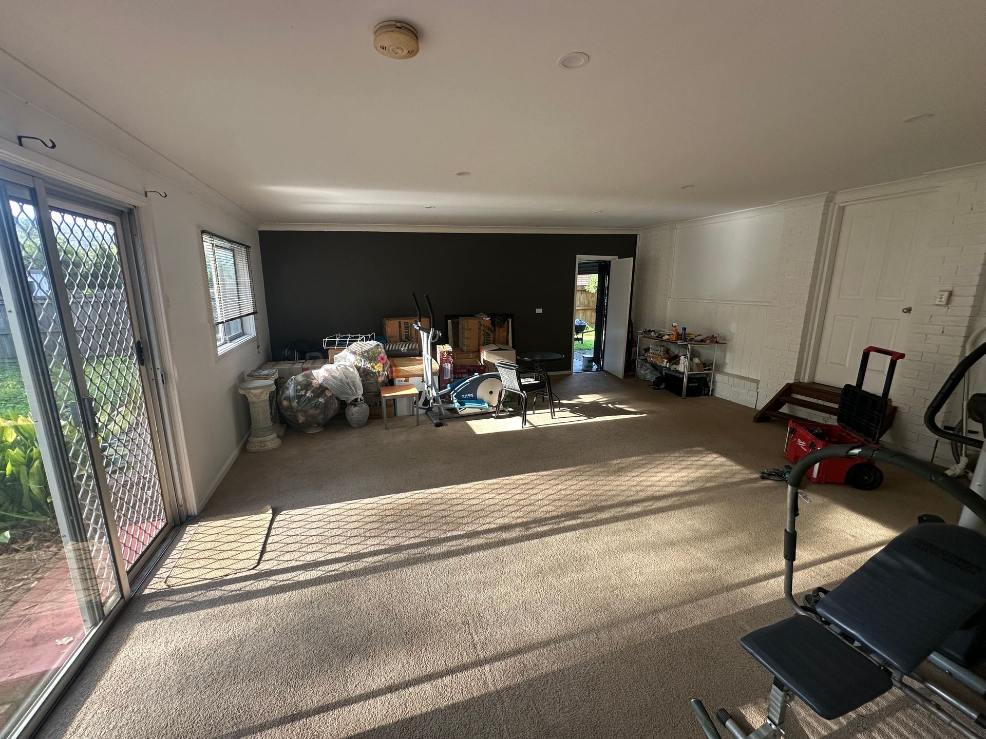A spacious room with carpeted floors, a sliding glass door, a dark accent wall, and some scattered gym and storage items.