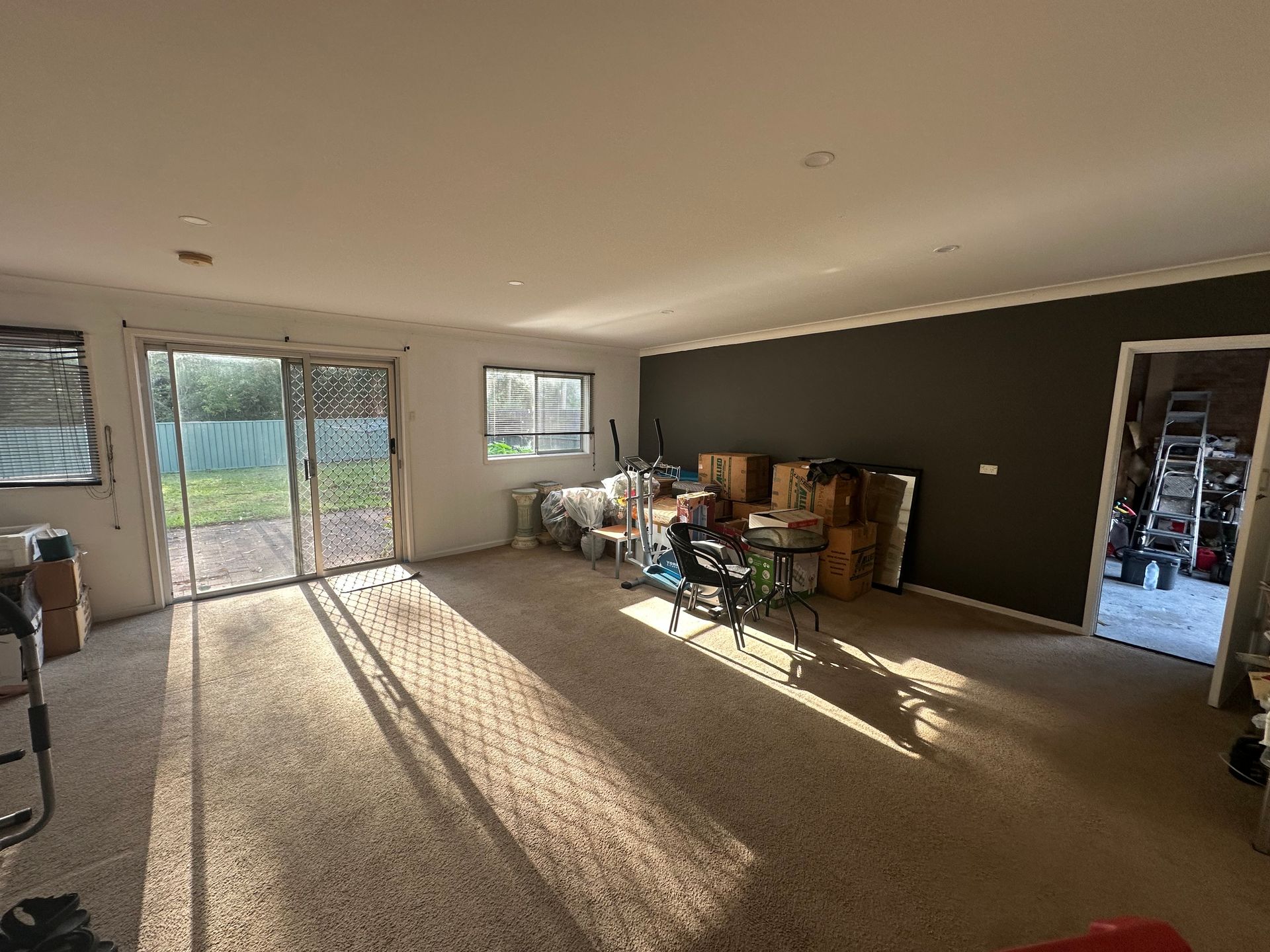 A sunlit living room with beige carpet, a sliding glass door, a dark gray accent wall, and moving boxes in the corner.