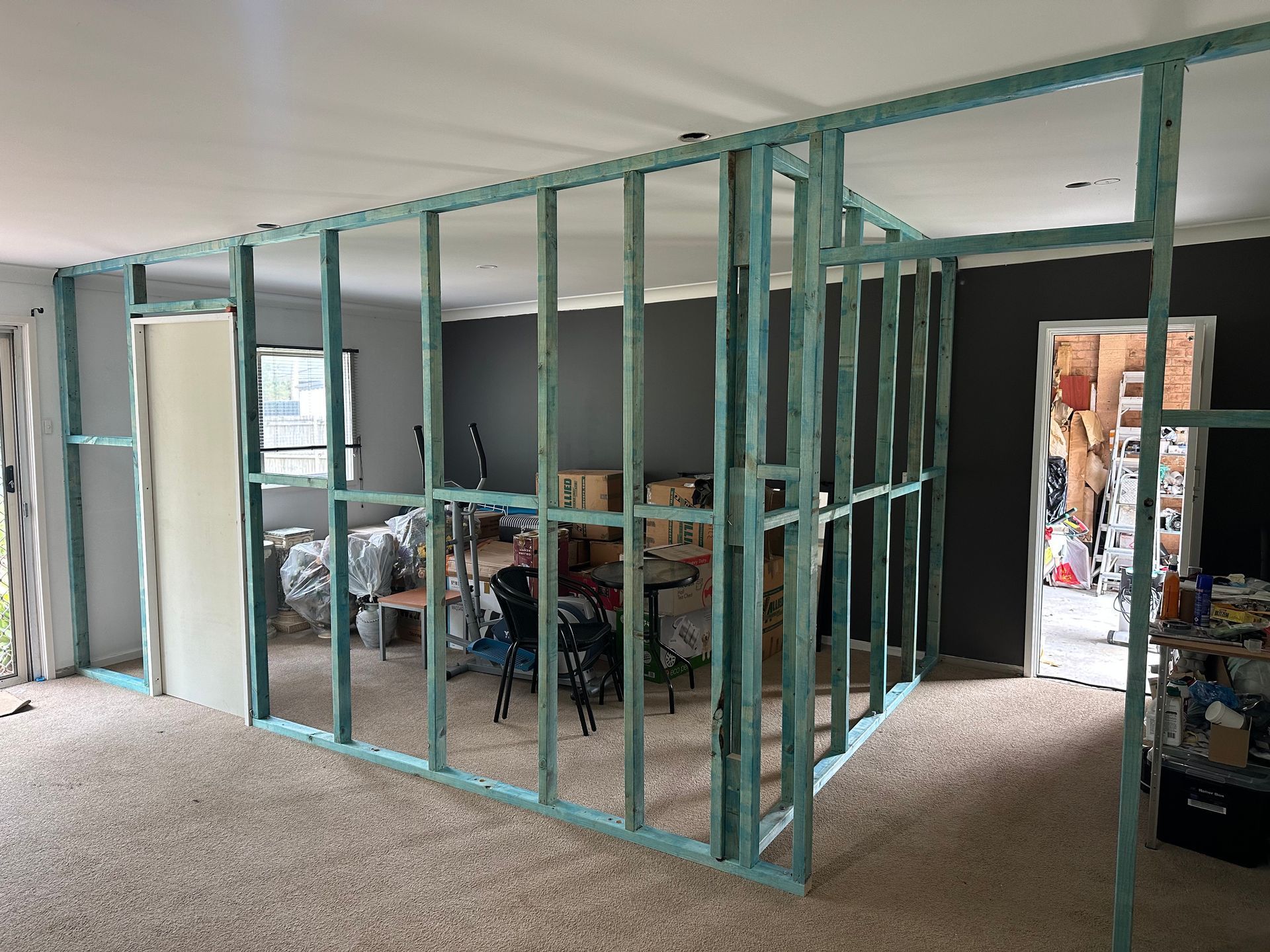 A room interior under renovation, featuring a partially built wooden frame wall structure atop a beige carpeted floor.