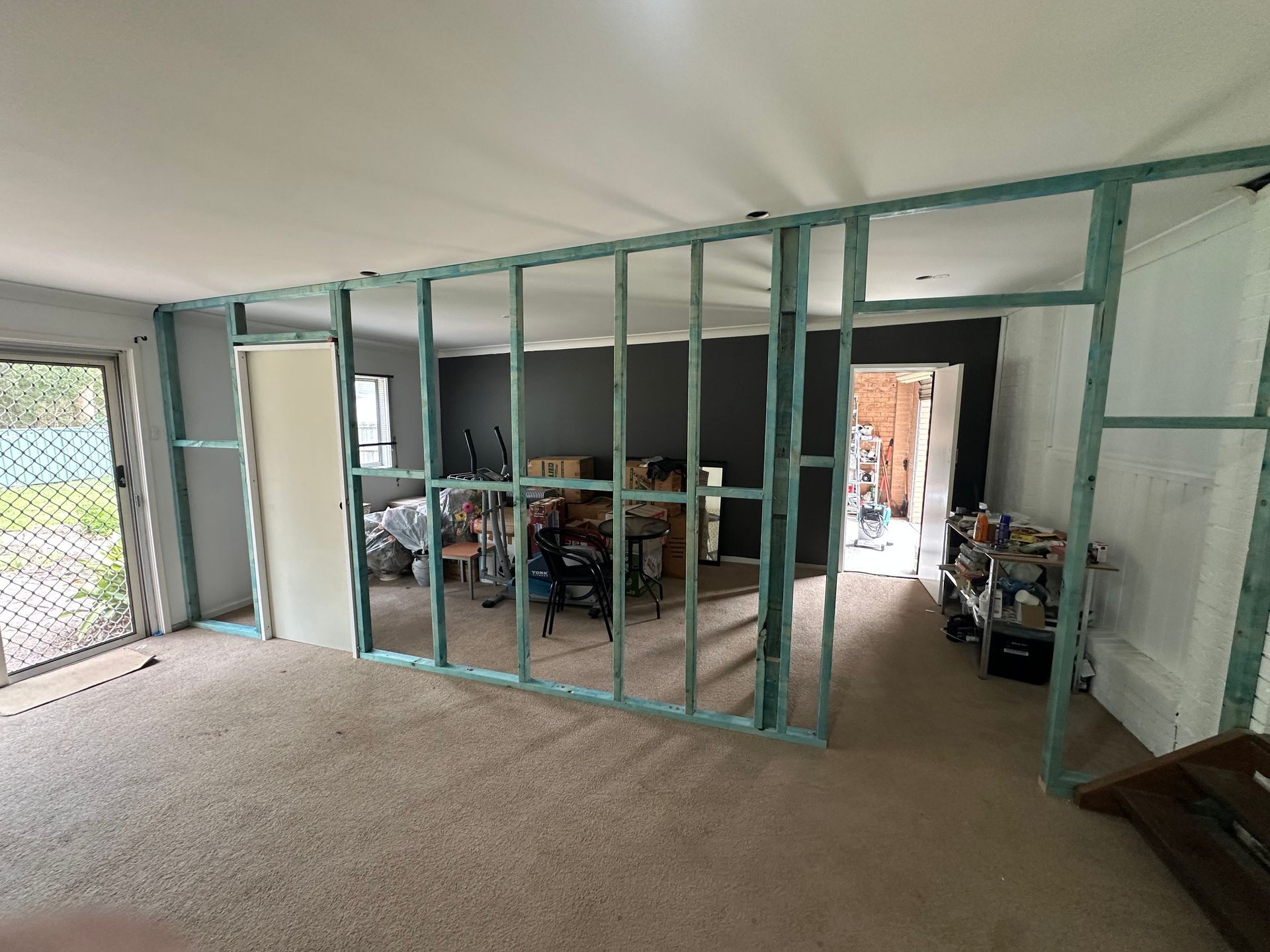 An unfinished wall frame made of light-blue timber studs divides an indoor room with beige carpet flooring.