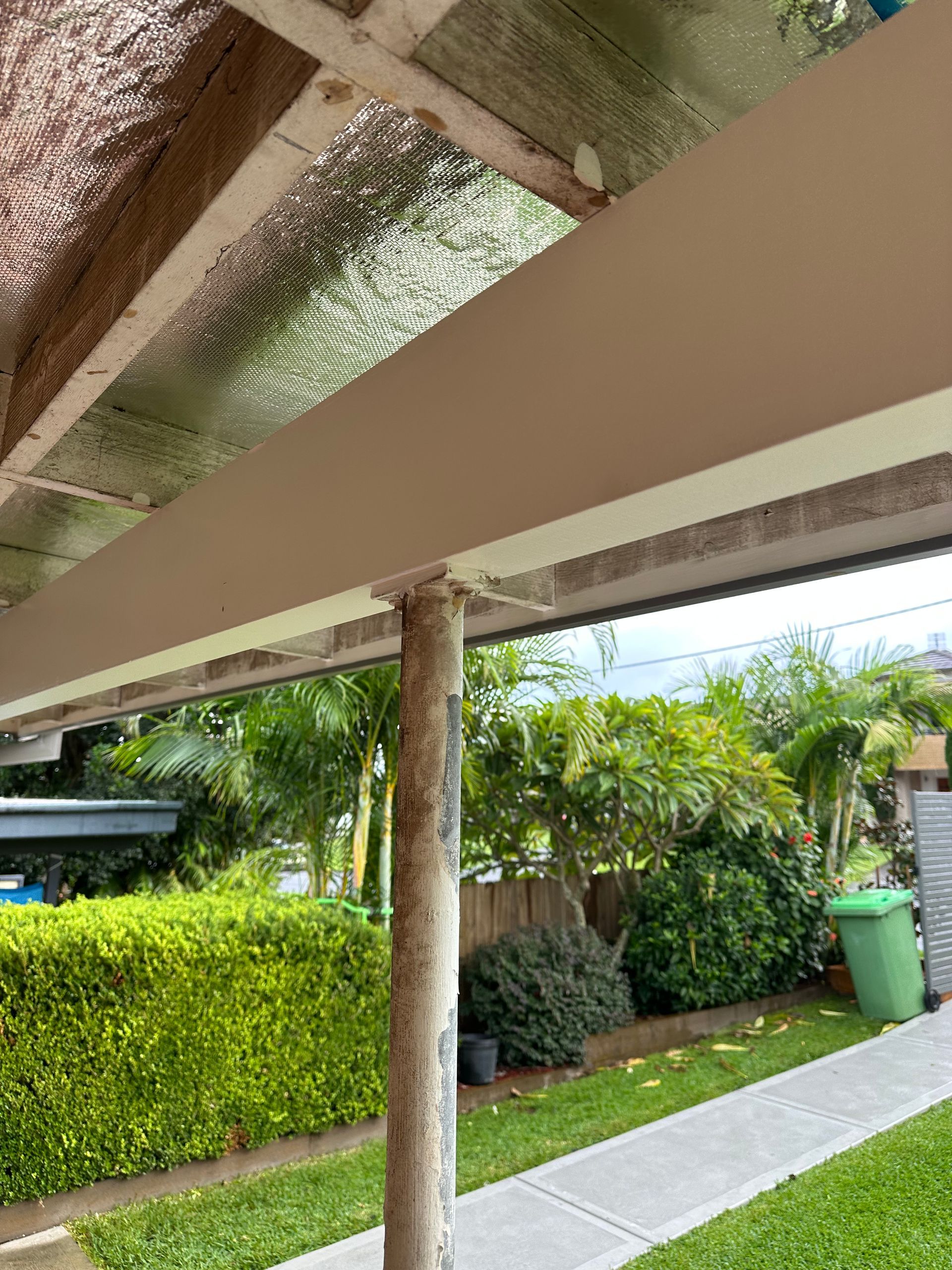 A weathered metal support post stands beneath a beige patio beam, overlooking a backyard with greenery and a walkway.