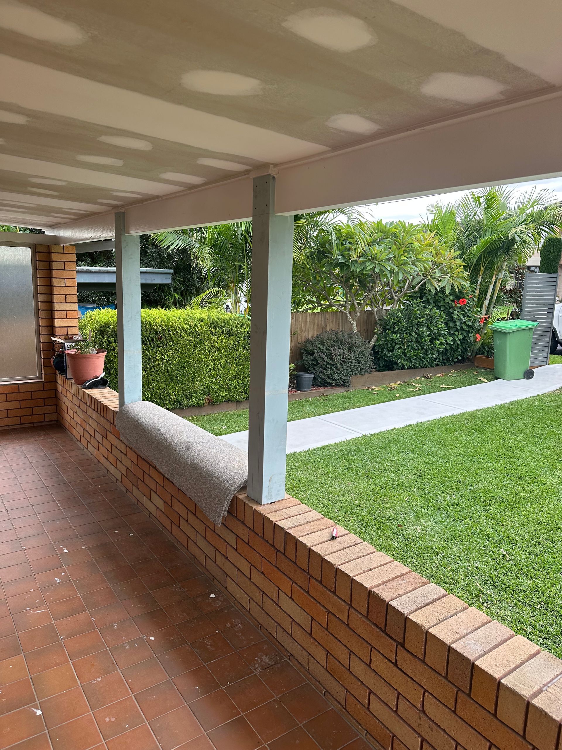 A brick patio bordered by a low brick wall, light blue wooden support posts, a grassy yard, and a house ceiling overhead.