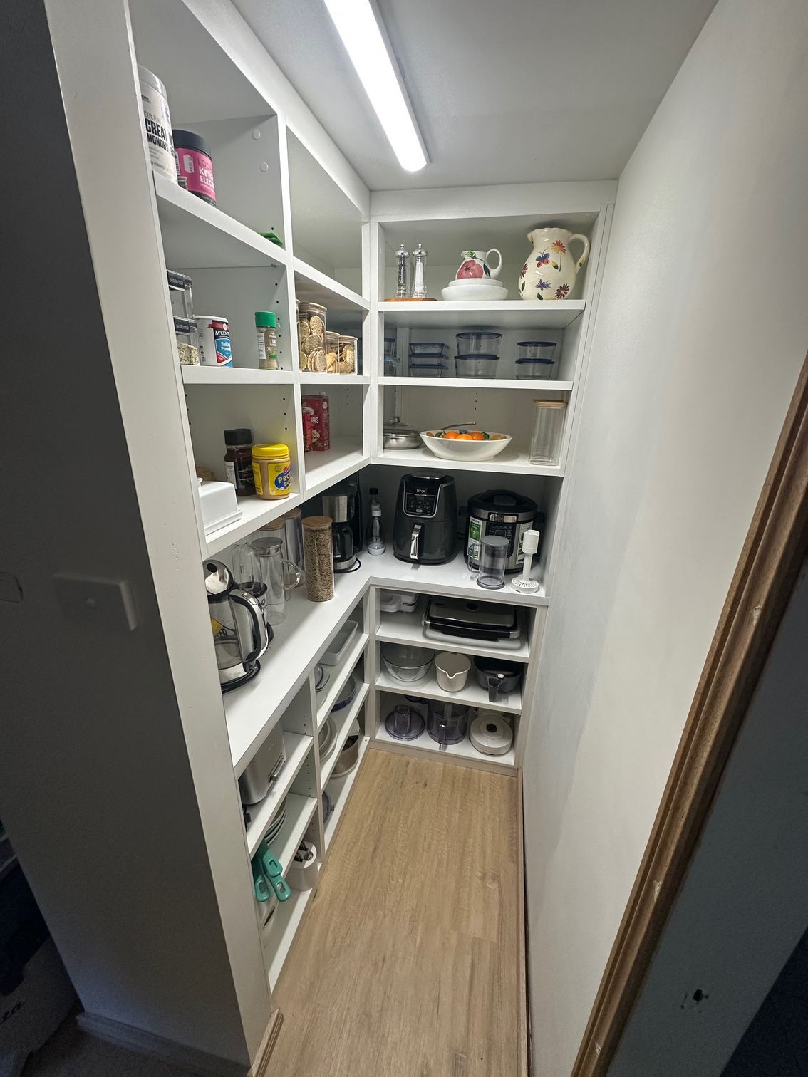 A walk-in white pantry with L-shaped shelves stocked with food items, kitchen appliances, and dishware on wood-look floors.