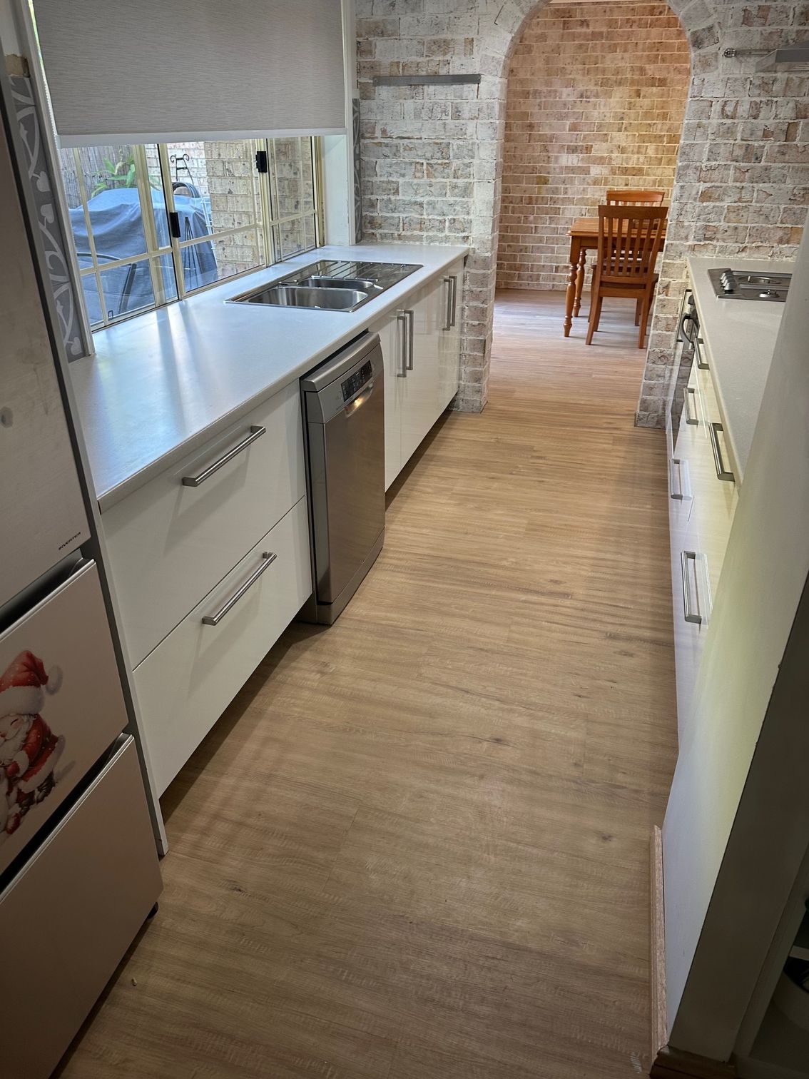 A white-cabinet kitchen with a stainless steel dishwasher, wood-look floors, and an arched brick wall leading to a dining area.