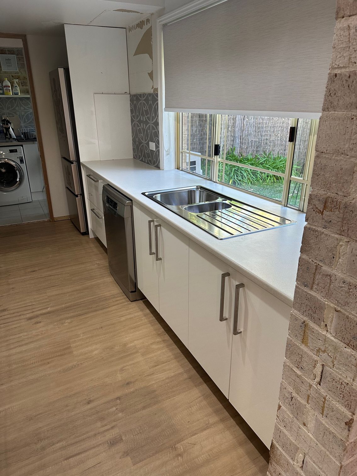 A modern kitchen featuring white cabinets, a stainless steel sink, a dishwasher, and a window with a grey roller blind.