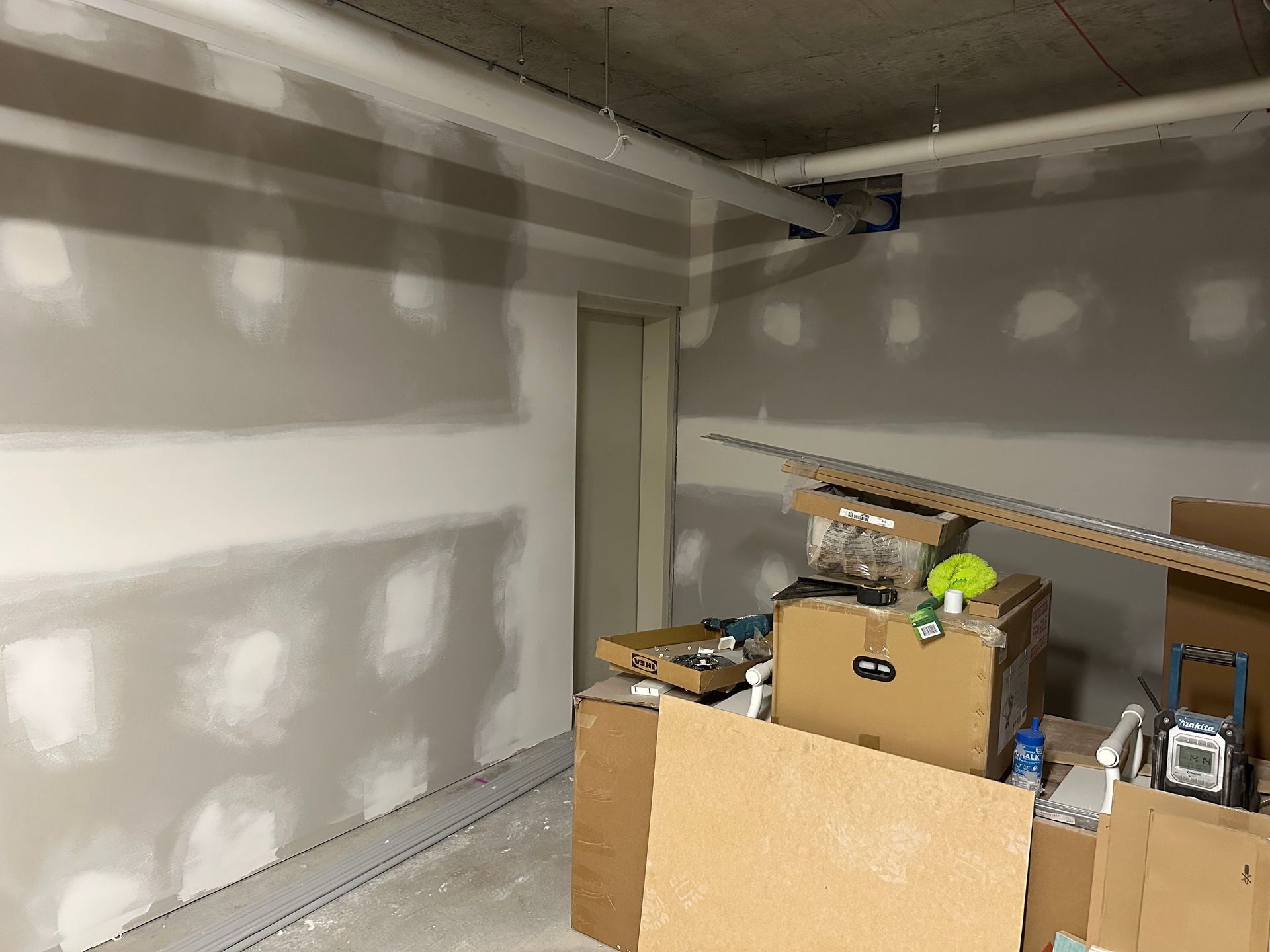 An interior room under construction with gray drywall featuring patched screw holes, exposed ceiling pipes, and boxes.