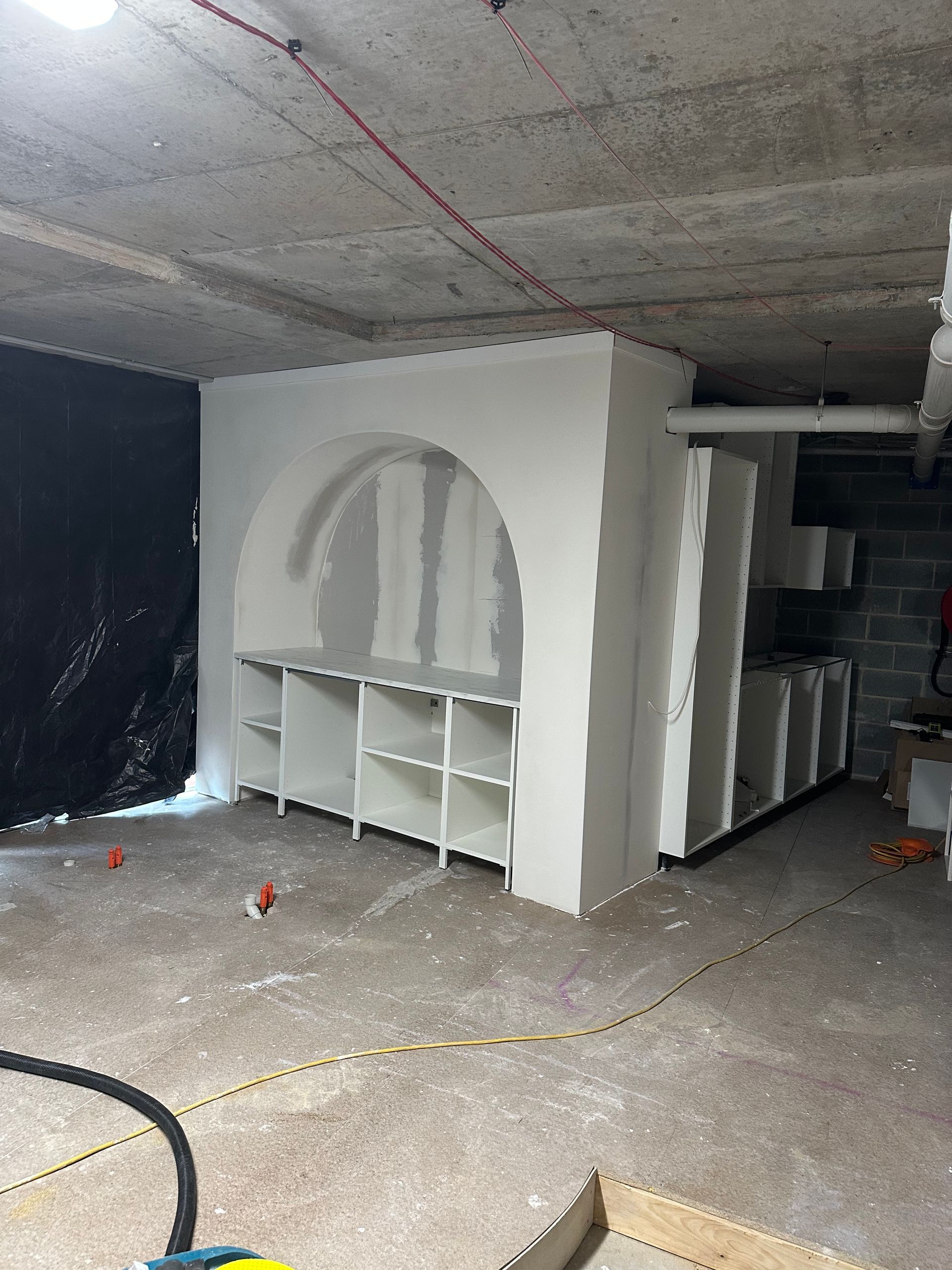 A basement room features a newly constructed white, arched drywall niche with built-in shelving undergoing renovation.