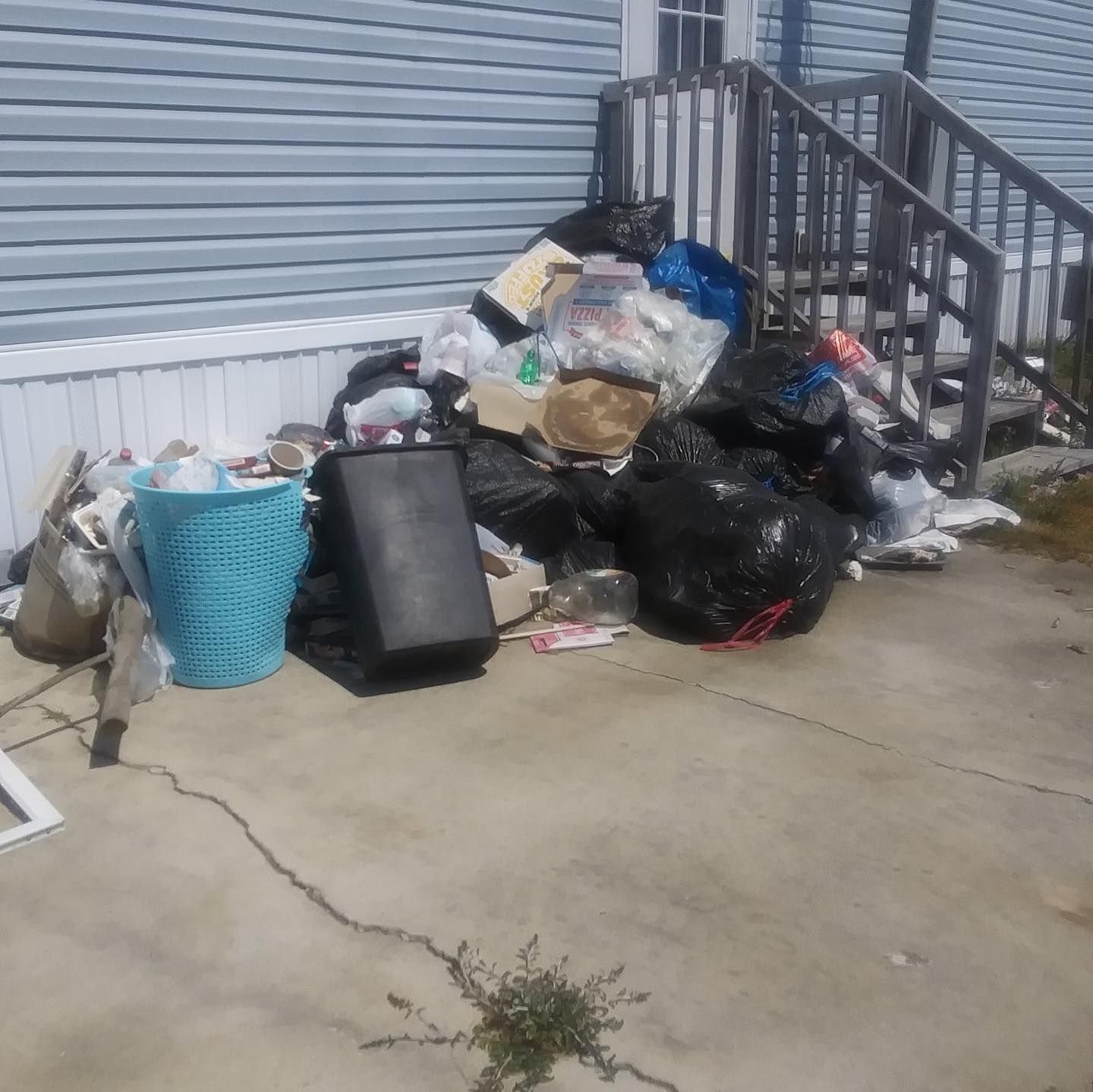 Pile of trash bags, containers, and debris on a concrete patio next to a light blue house with stairs.