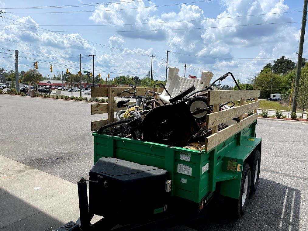 Green trailer loaded with yard waste, parked on a sunny street.