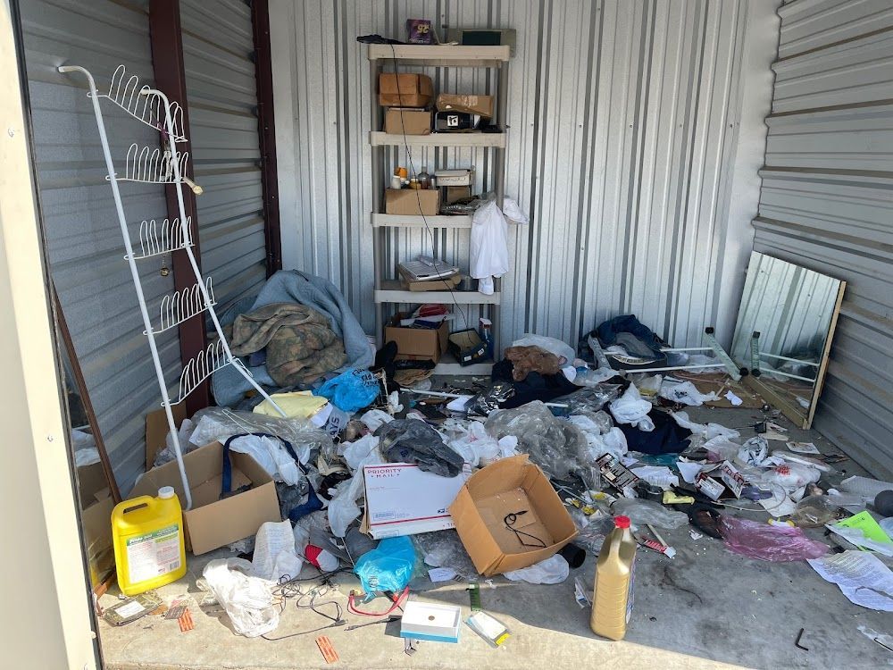 Cluttered storage unit: shelf, clothes, trash, boxes, and liquid container.