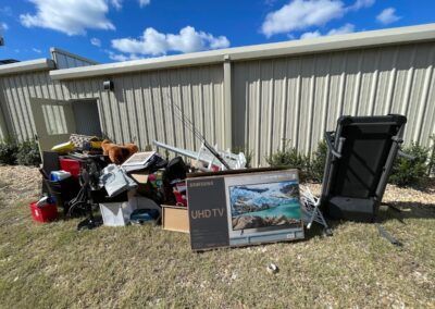 Pile of discarded items, including a TV box and a treadmill, by a building on grass.