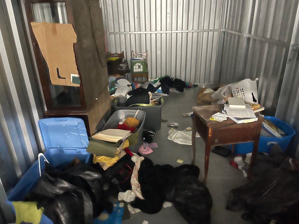 Cluttered storage unit with boxes, furniture, and various items scattered on the floor.