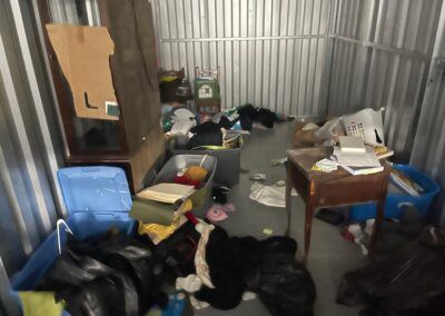 Cluttered storage unit: various items scattered on floor, including boxes, furniture, and trash bags.