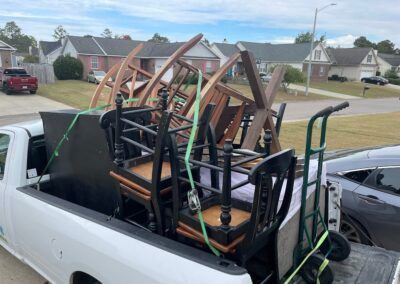White pickup truck bed loaded with chairs and a table. Green straps secure the furniture in a residential area.
