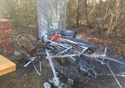 Pile of discarded metal, trash, and debris in a wooded area, including a lawnmower.