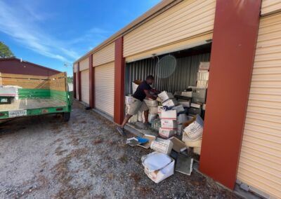 Man unloads storage unit with boxes. Beige garage door, red trim. Green truck waits outside.