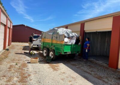 People loading a green trailer with items from a storage unit under a blue sky.