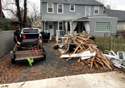 Man loading debris into a trailer next to a gray house with a pile of wood.